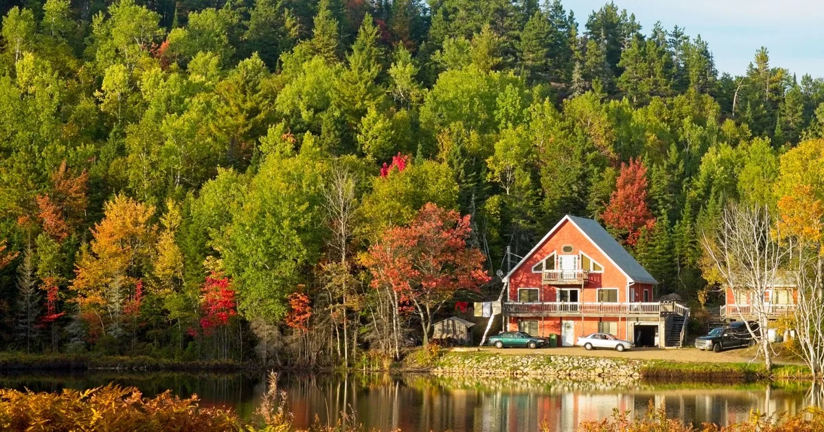 A red two-story lake house on a full concrete foundation with a gravel drive, set against pine and fall-color hills beside a calm lake, for a guide to financing a Maine camp or lake house