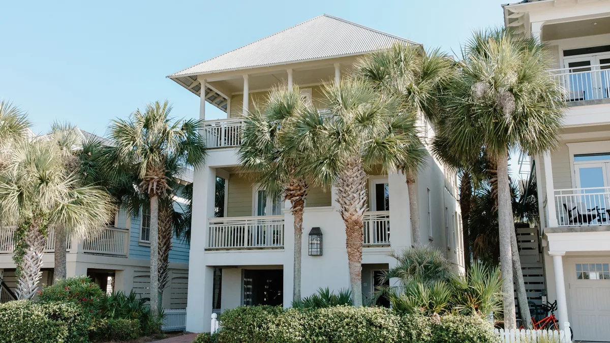 Raised white coastal home with deep porches, palm trees and a picket fence under a clear blue sky