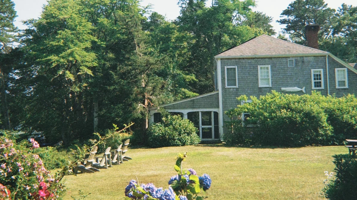 Gray shingled two story home with a screened porch, a wide summer lawn, Adirondack chairs and blue hydrangeas
