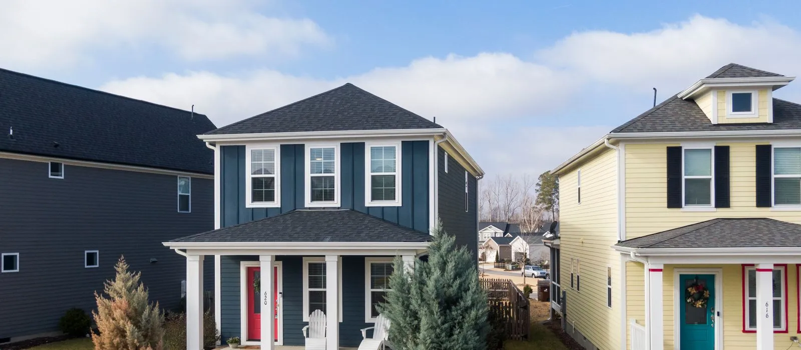 Two story houses with front porches side by side on a residential street, one navy blue and one yellow, under a partly cloudy sky