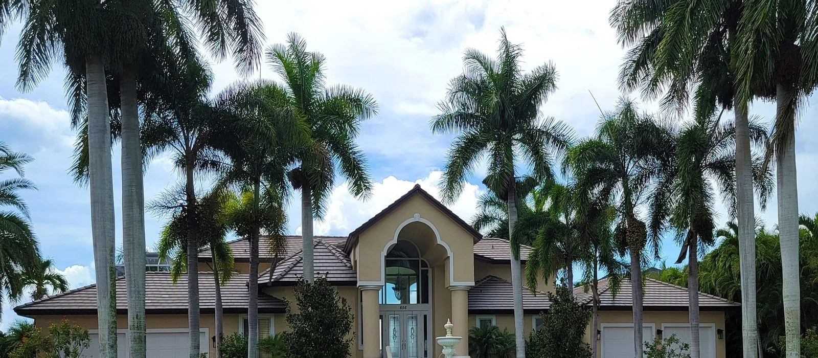 Tan single story Florida home with a tile roof, arched entry and fountain, framed by tall palm trees on a green lawn